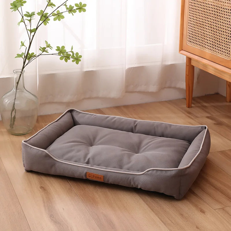 A gray waterproof Oxford dog bed placed on a wooden floor against a wall, with a plant vase in the background.