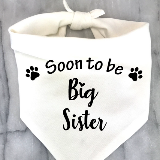 A white pet bandana with the text 'Soon to be Big Sister' printed in black, featuring paw prints as decorative elements.