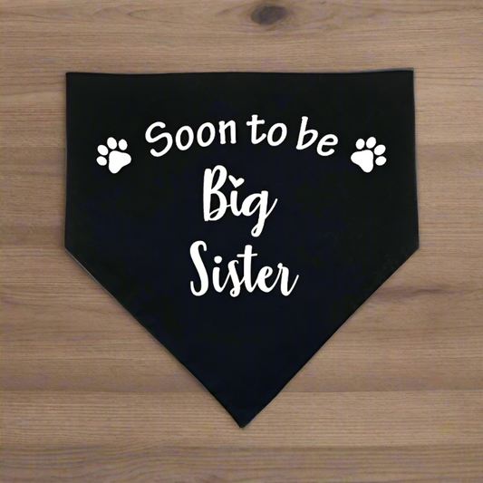 A black pet bandana with white text 'Soon to be Big Sister' and white paw prints.